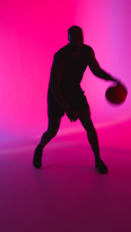 Vertikale-Videostudio-Silhouette-Eines-Männlichen-Basketballspielers,-Der-In-Rael-Zeit-Einen-Ball-Vor-Rosa-Und-Blau-Beleuchtetem-Hintergrund-Dribbelt-Und-Hüpft